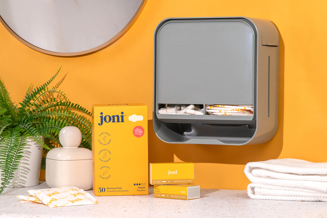 joni Model A dispenser on an orange wall beside tampon and pad boxes and folded towels