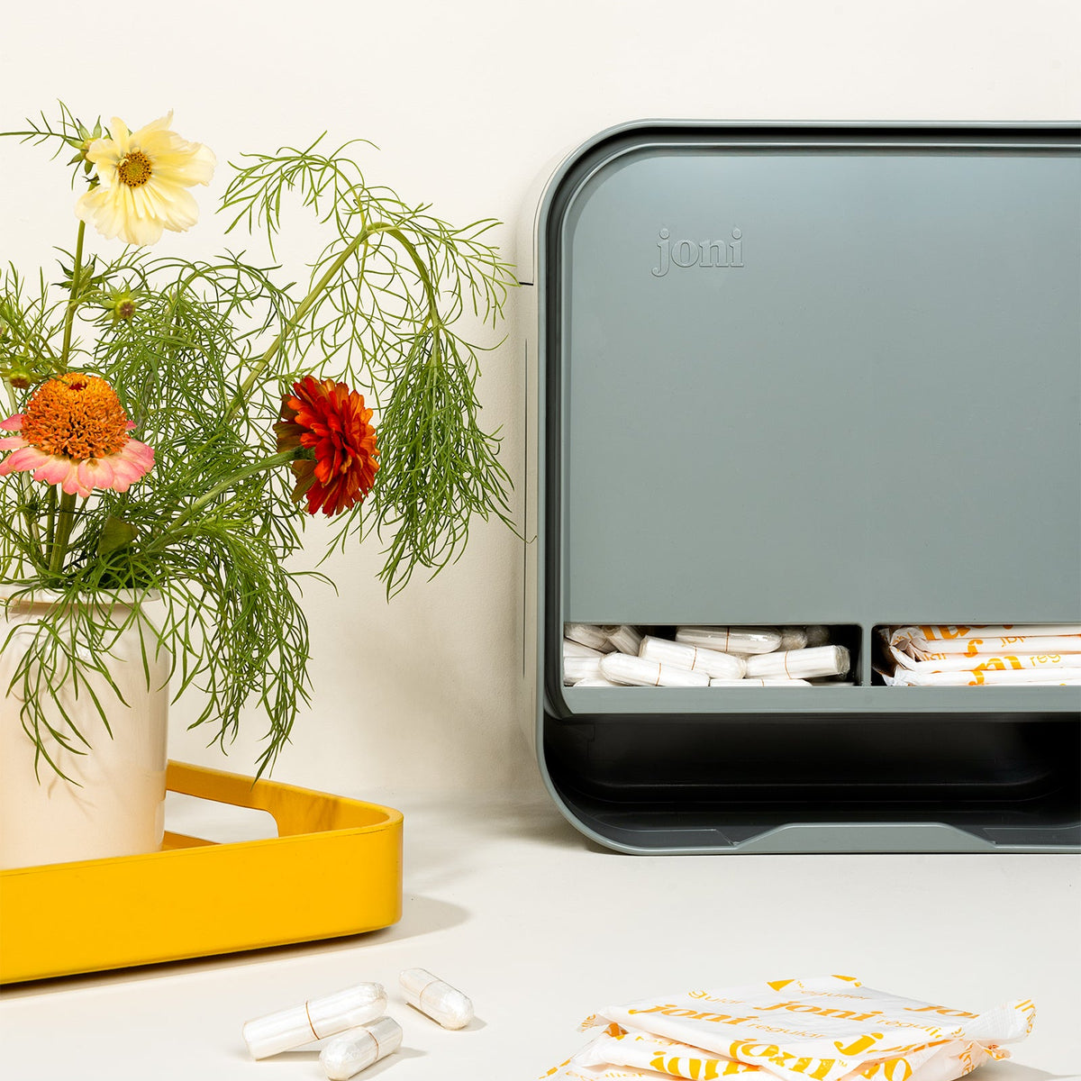 A joni open period care dispenser pill organizer box in a muted gray-green color, styled on a white surface next to a yellow tray with flowers and tampons.