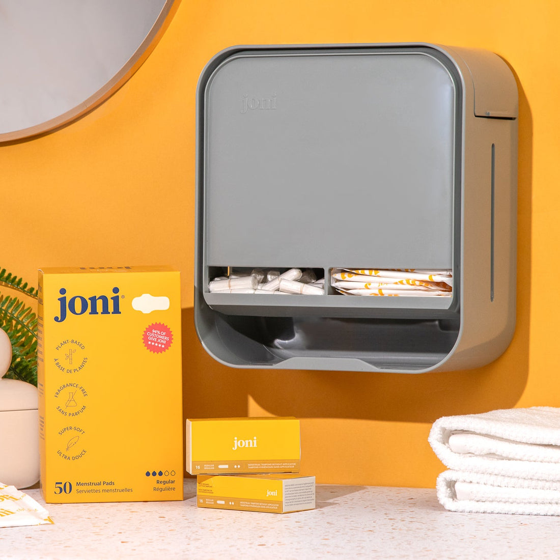 A wall-mounted dispenser holding Joni pads and tampons  on a countertop against a warm yellow wall. A plant, ceramic jar, folded towels, and part of a mirror are visible.