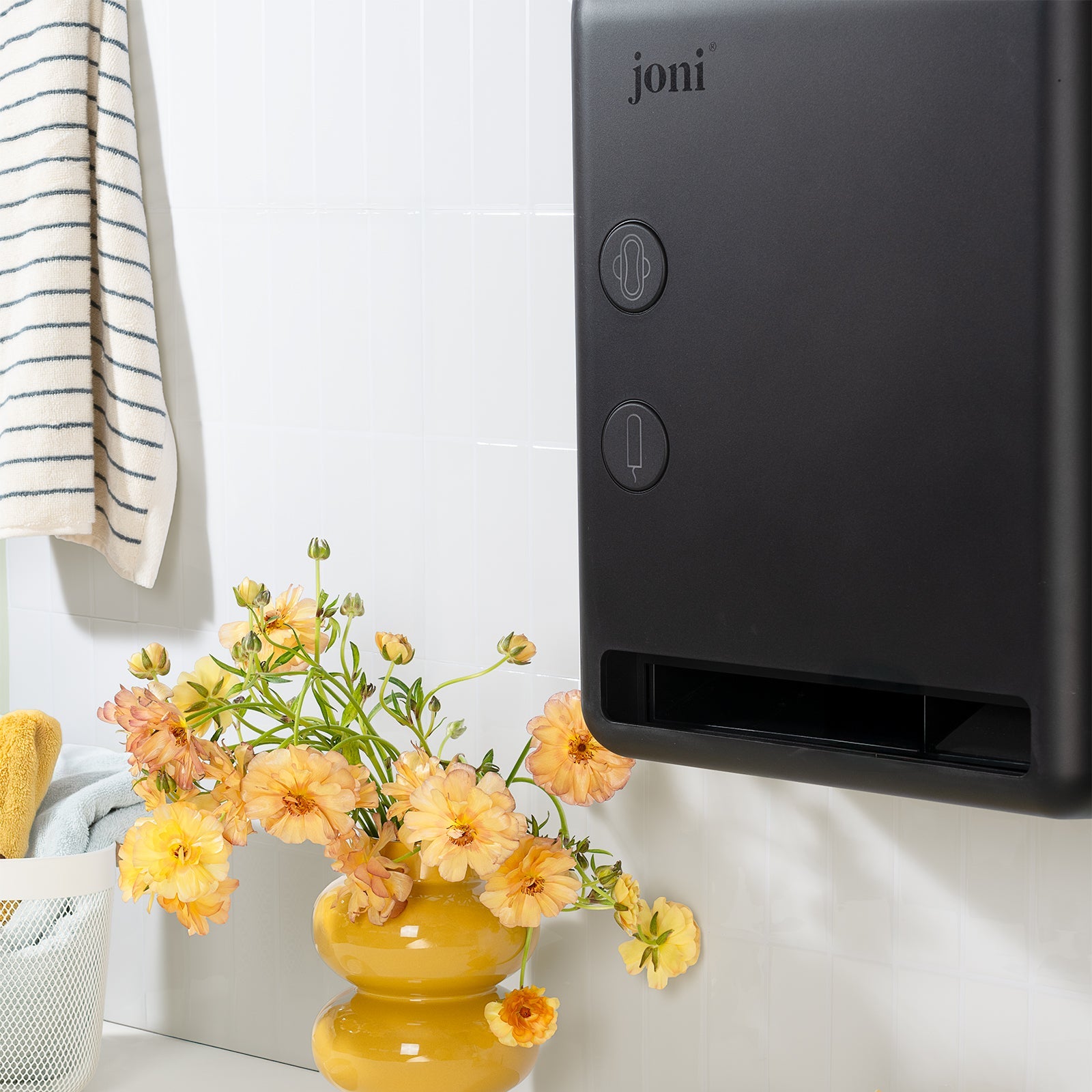 A wall-mounted joni smart period care dispenser in a bright bathroom setting, shown next to a yellow vase with flowers and two yellow Joni paper towel packs on a white countertop.