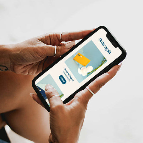 A close-up of a person holding a smartphone with both hands, showing the 'Orderagain' mobile application on the screen against a soft, light gray background.