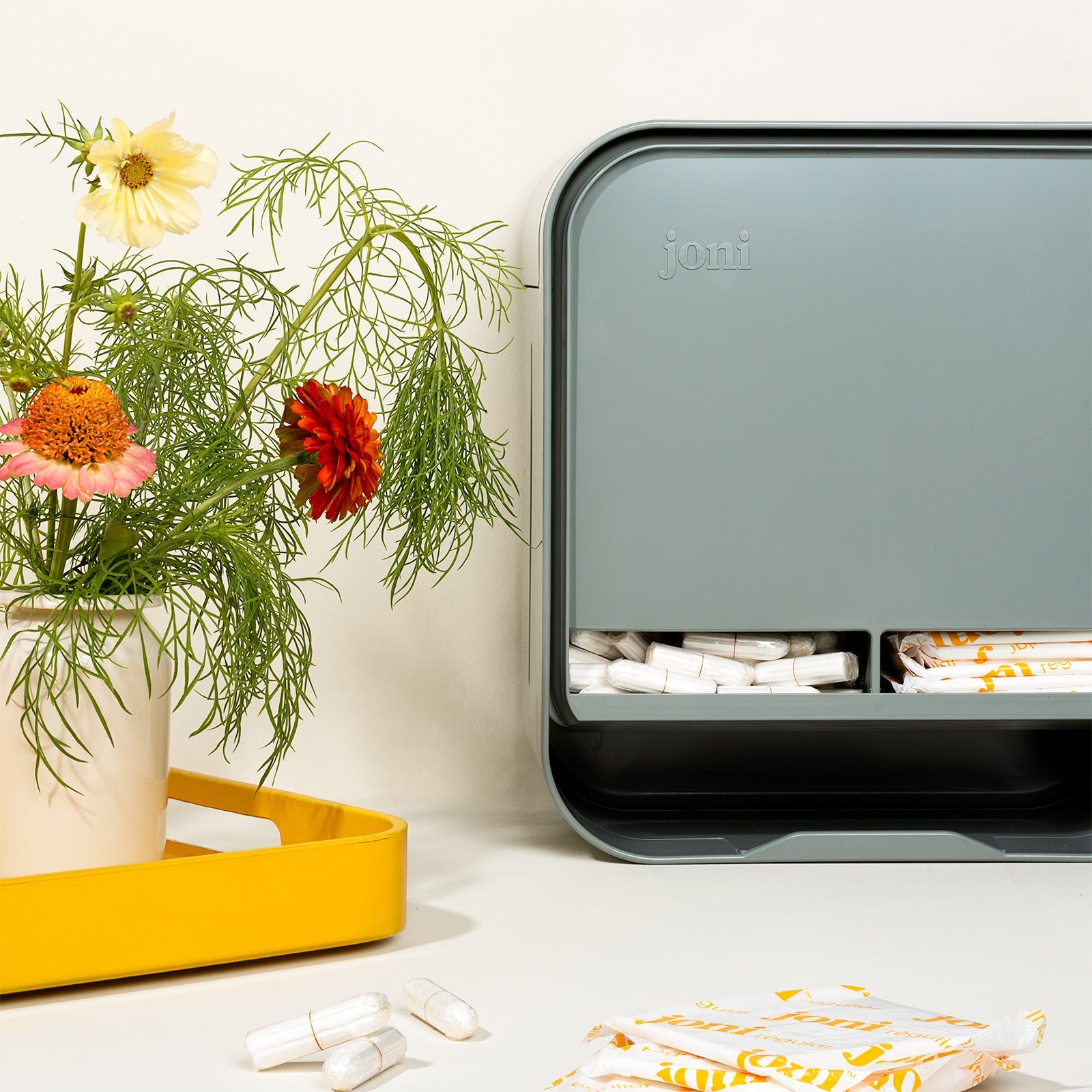 A joni period care open dispenser organizer box in a muted gray-green color, styled on a white surface next to a yellow tray with flowers and tampons.