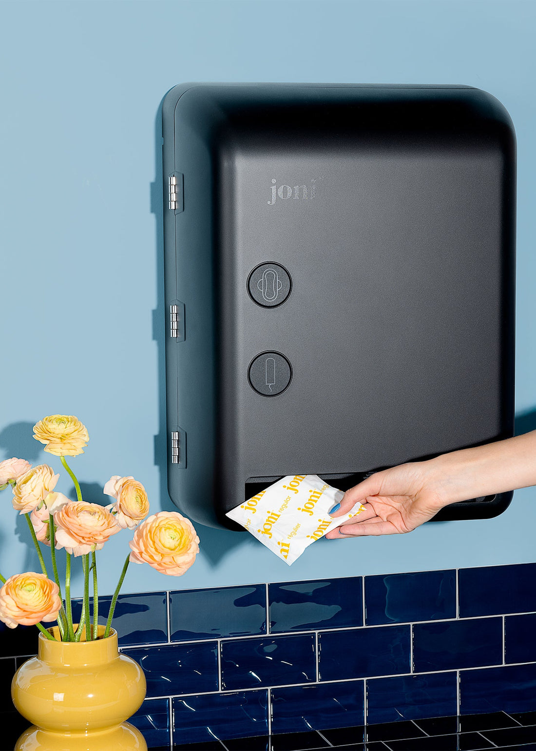 Hand placing a packet into a joni model v smart period care dispenser against a blue tiled wall with flowers.