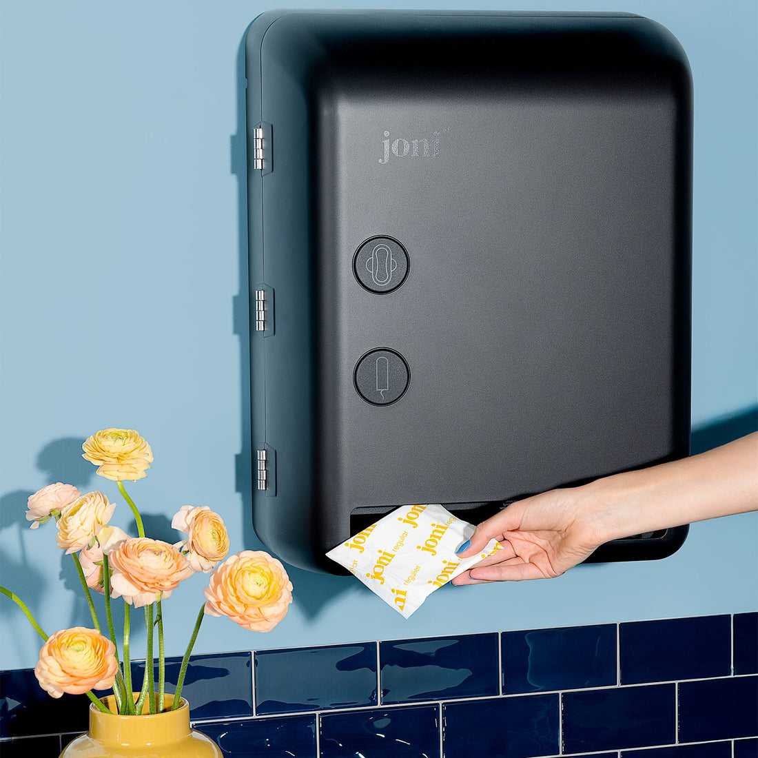 Hand placing a packet into a joni model v period care smart dispenser against a blue tiled wall with flowers.