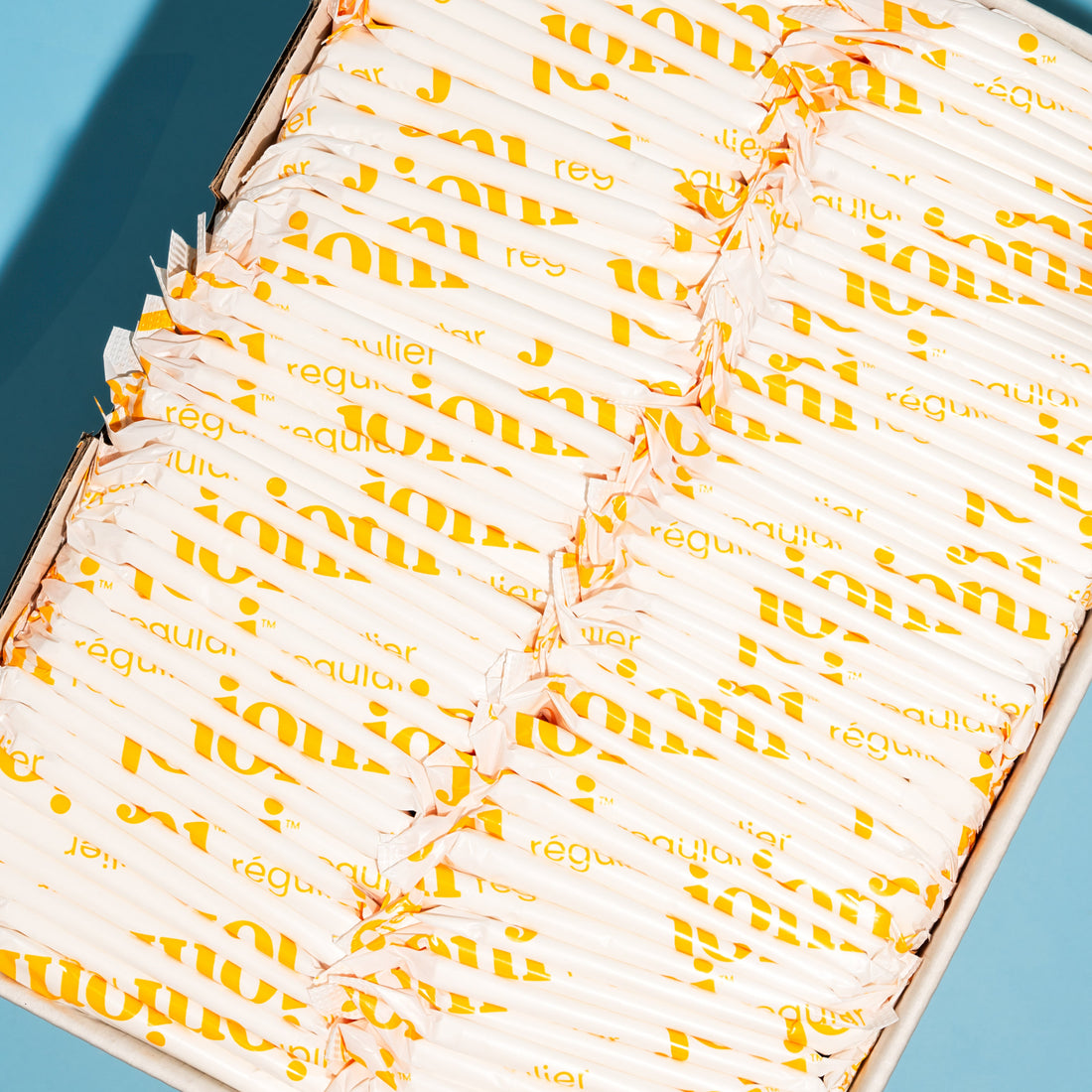 A box filled with individually wrapped joni regular pads printed photographed from above on a bright blue background.