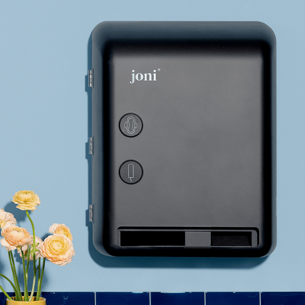 joni Model V electronic period care dispenser on a blue wall with blue tile and a yellow flower vase